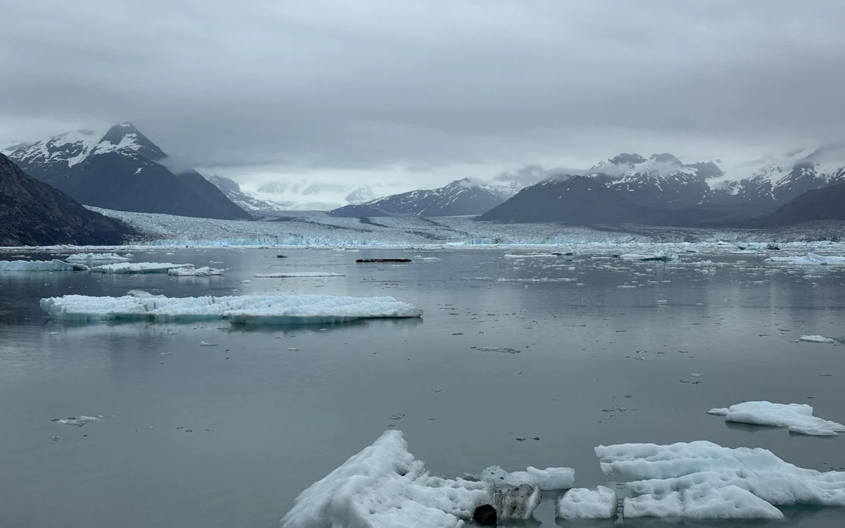 Passengers in windproof jackets on the open deck of a Kenai Fjords cruise boat with spray and mountains behind